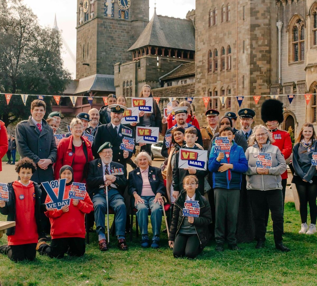 A group of adults and school children holding up VE Mail signs. There are two World War II veterans in the middle of the group